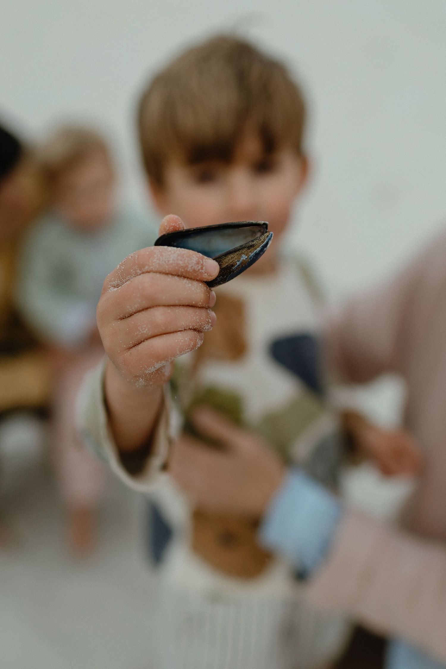 A young boy holds up a single mussel shell toward the camera while family members sit blurred in the background.