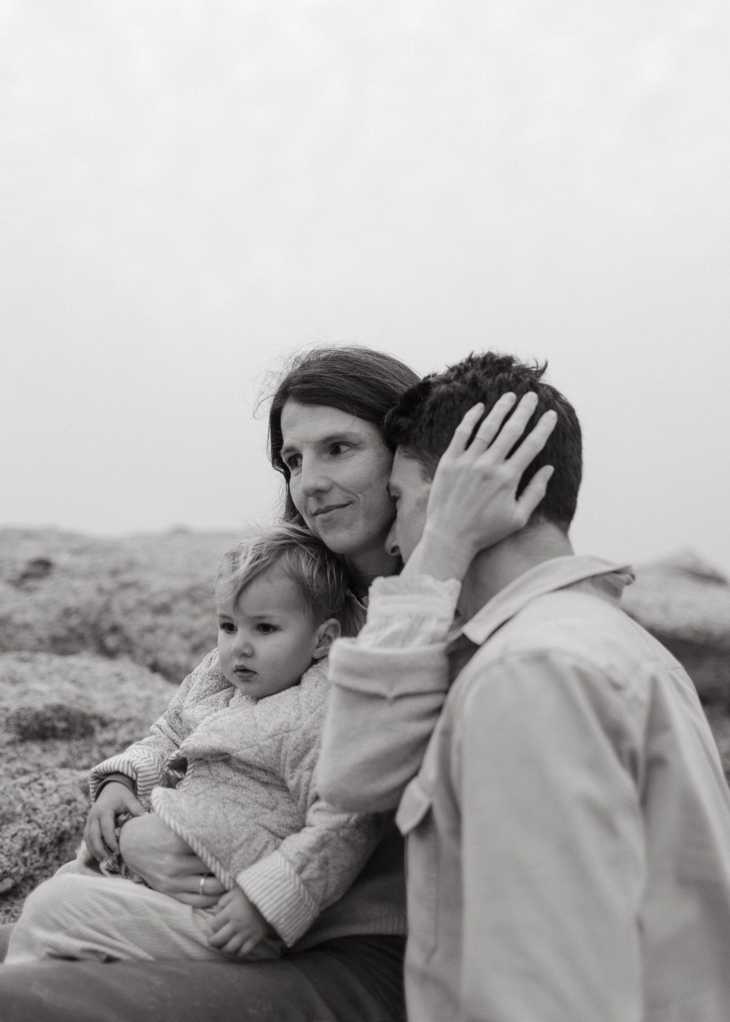 Black and white image of a mother holding her toddler while a father rests his head against her on a misty coast.