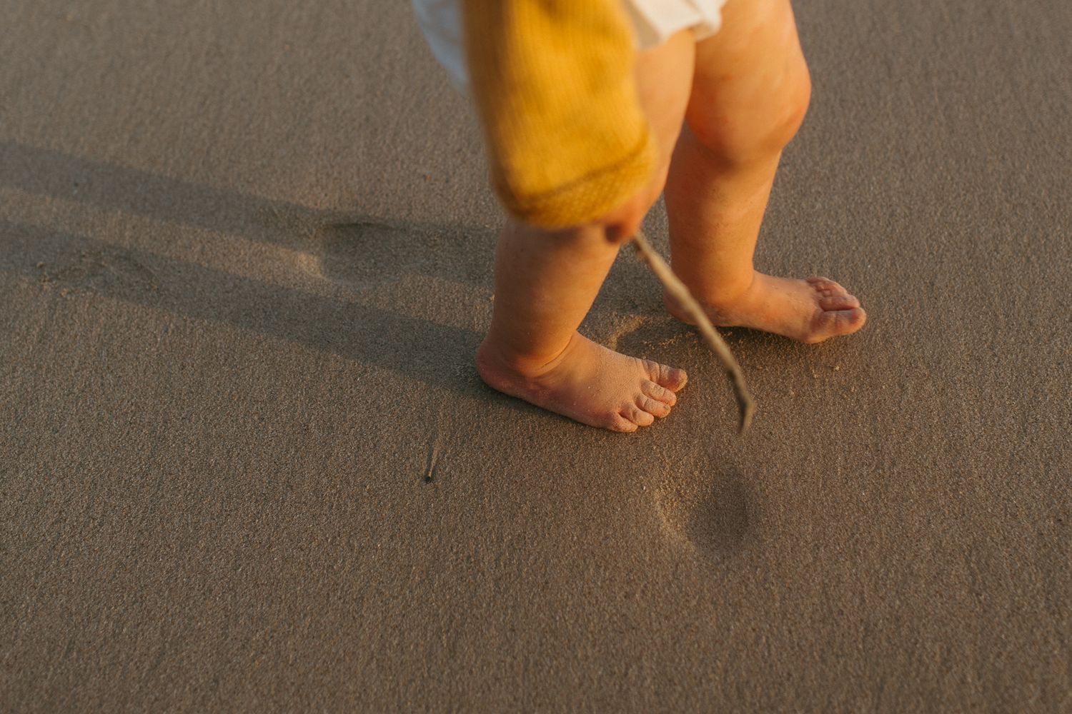 Toddler's bare feet and legs stepping on smooth wet sand, wearing a yellow cardigan and holding a small stick.