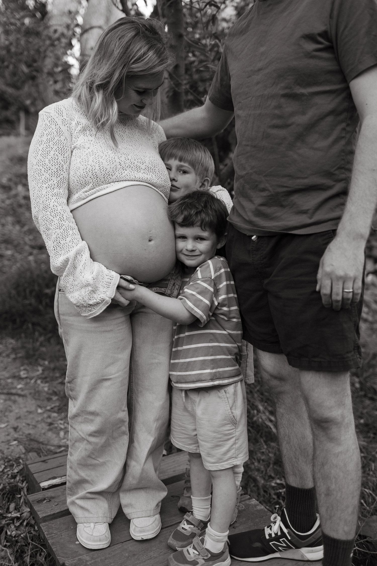 A pregnant mother and father stand with two young sons who hug her baby bump in a black and white photo.