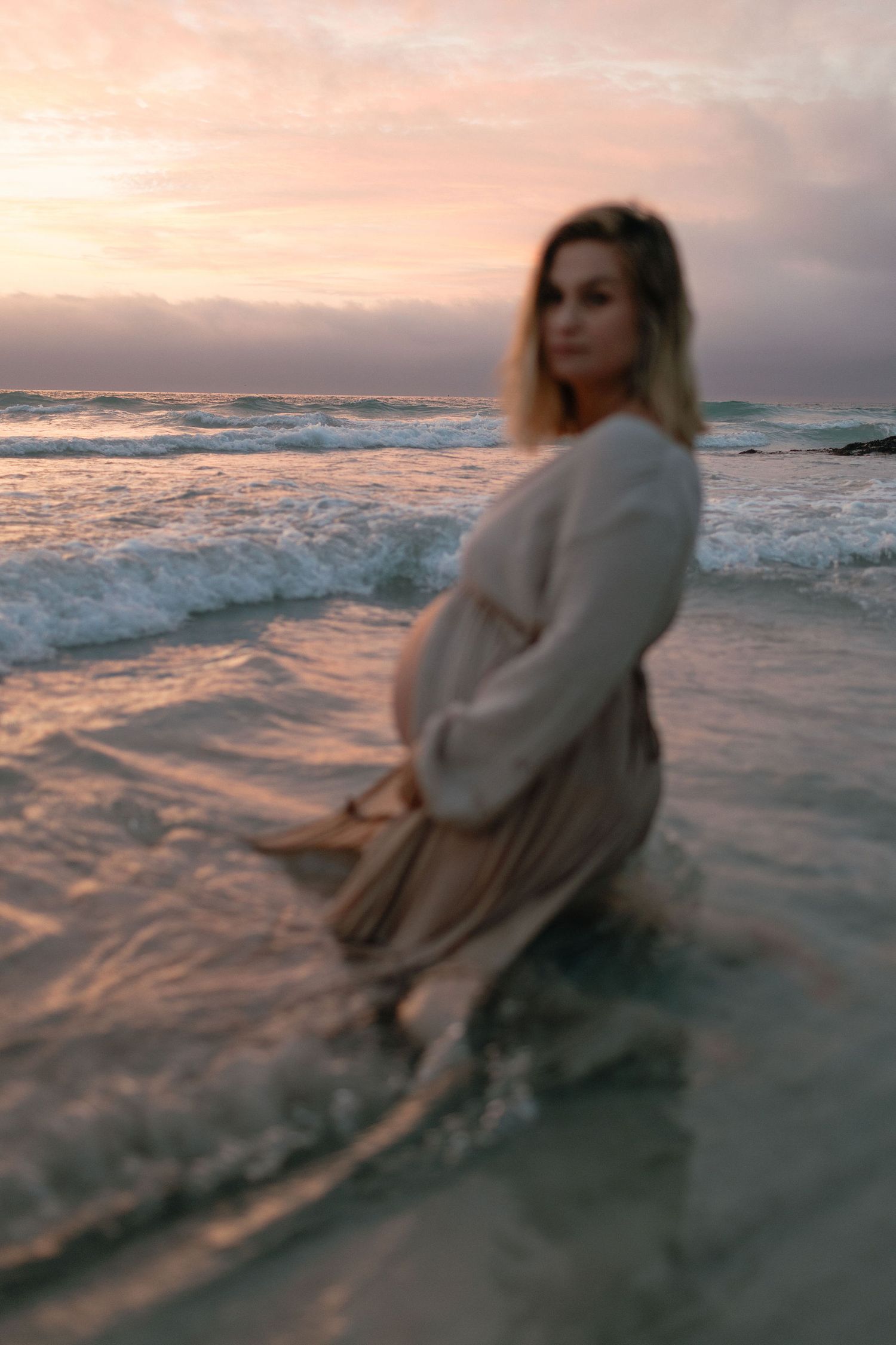 A blurry, dreamy photo of a pregnant woman in a beige dress standing in shallow ocean waves at a pink sunset.