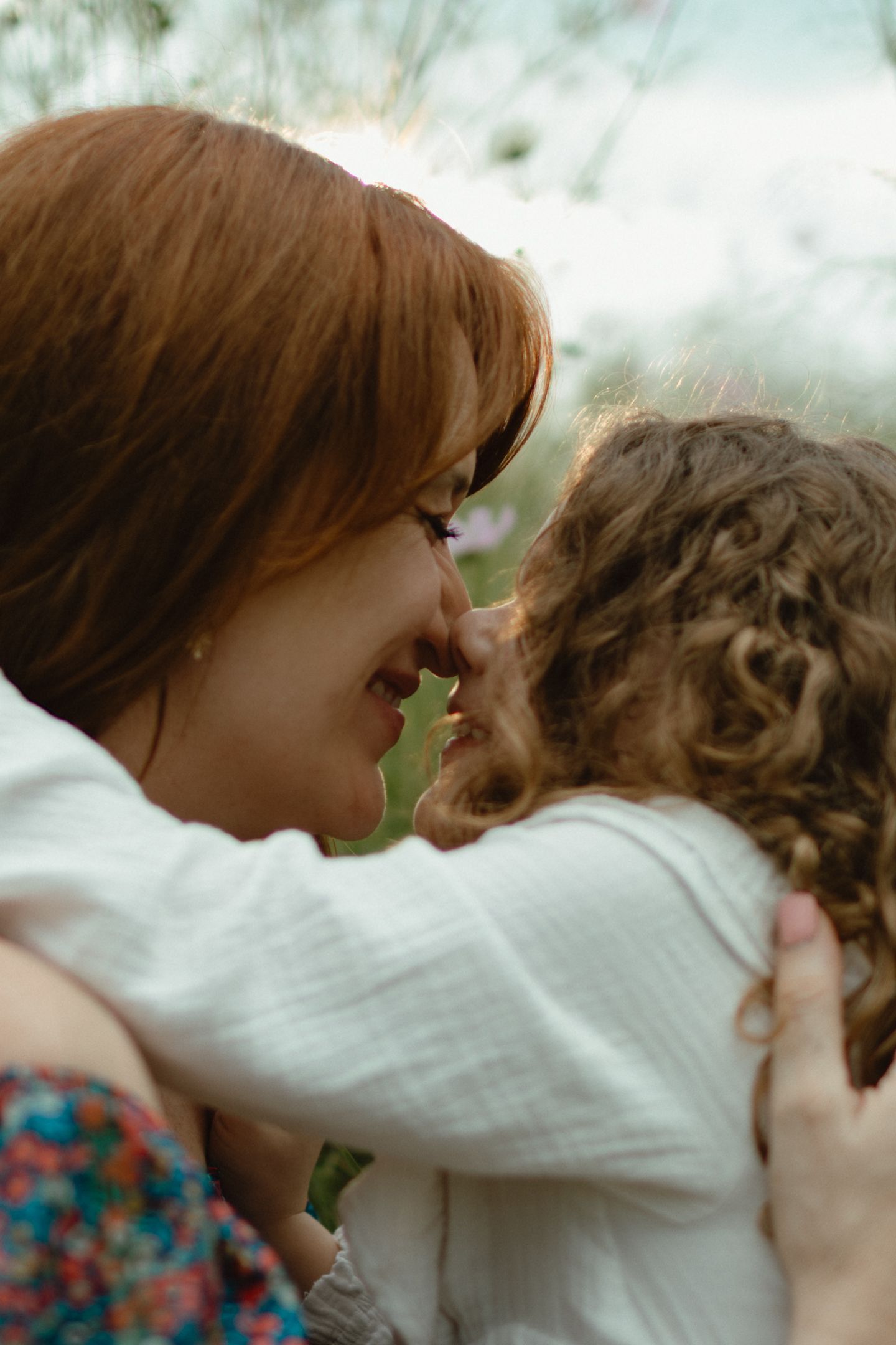 A smiling red-haired mother touches noses with her curly-haired child in a tender embrace outdoors.