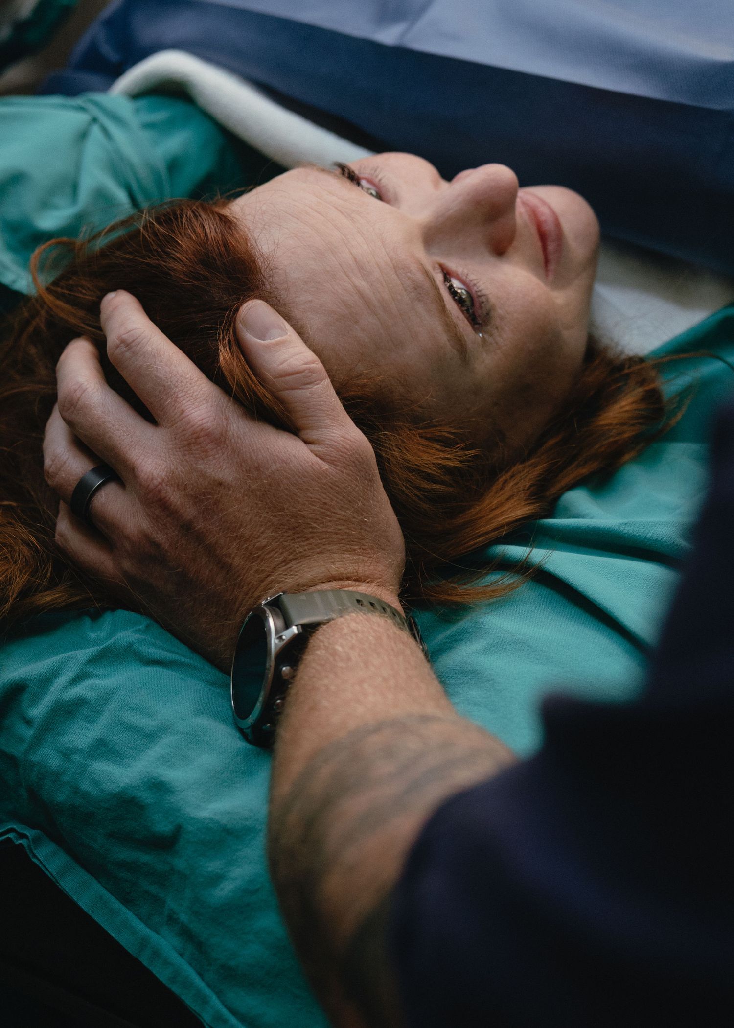 A red-haired woman lies on a hospital table in teal drapes while a partner gently cradles her head during labor and delivery.