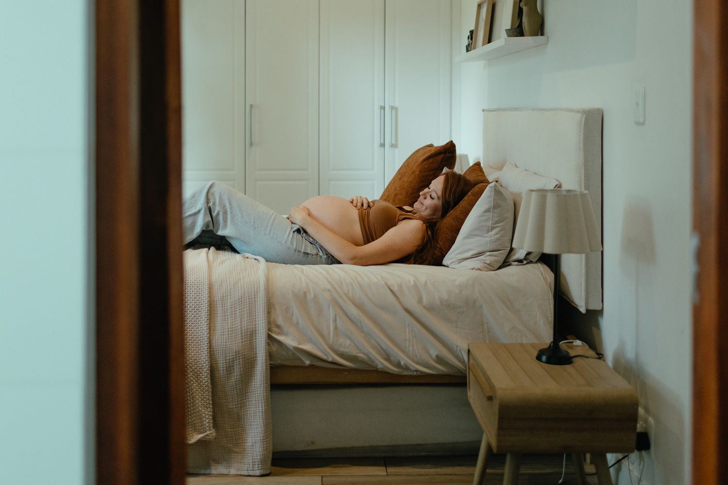 A pregnant woman lies on her side on a bed with pillows in a calm, softly lit bedroom seen through a doorway.