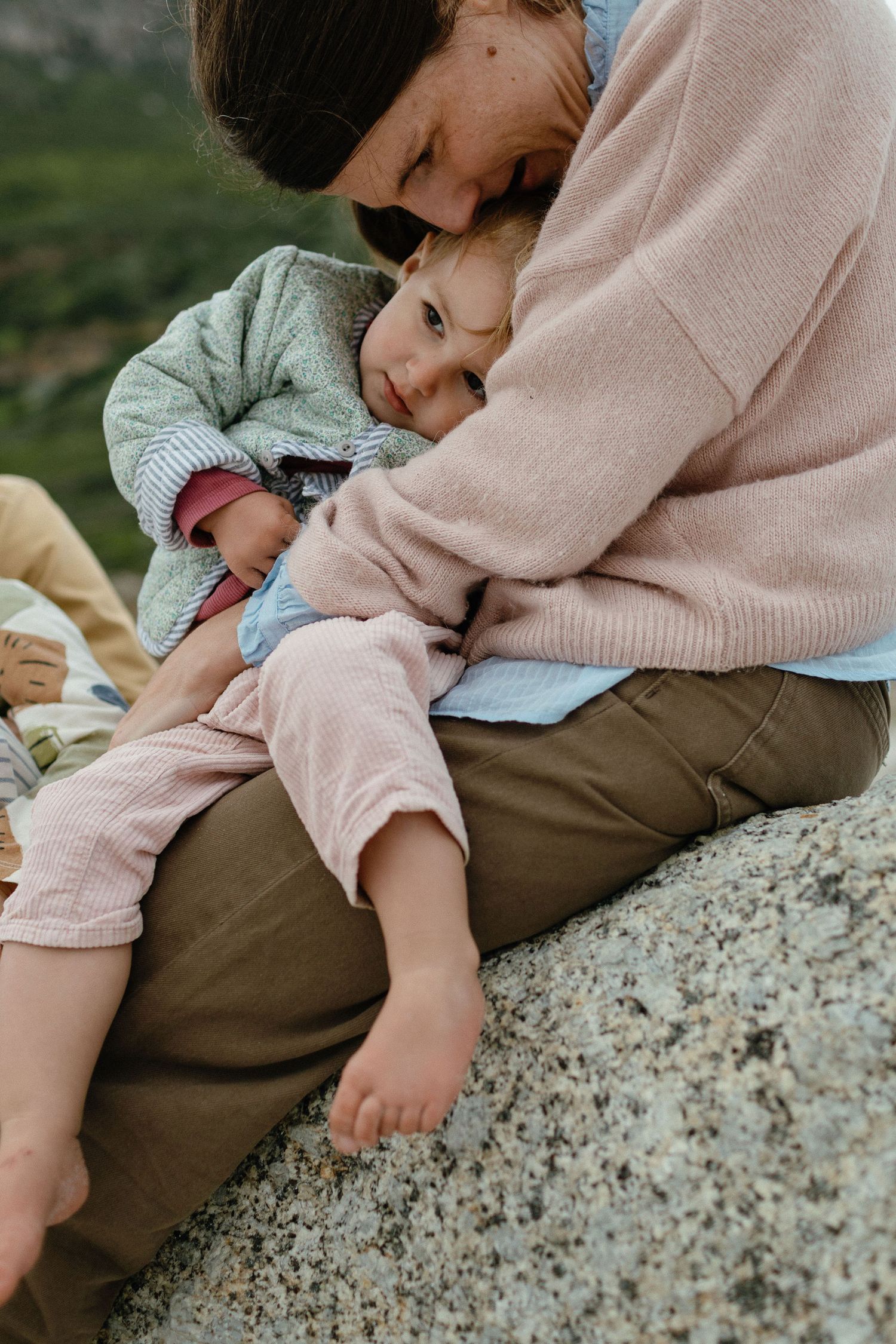 A mother in a pink sweater cuddles a barefoot toddler resting against her as they sit on a granite rock outdoors.