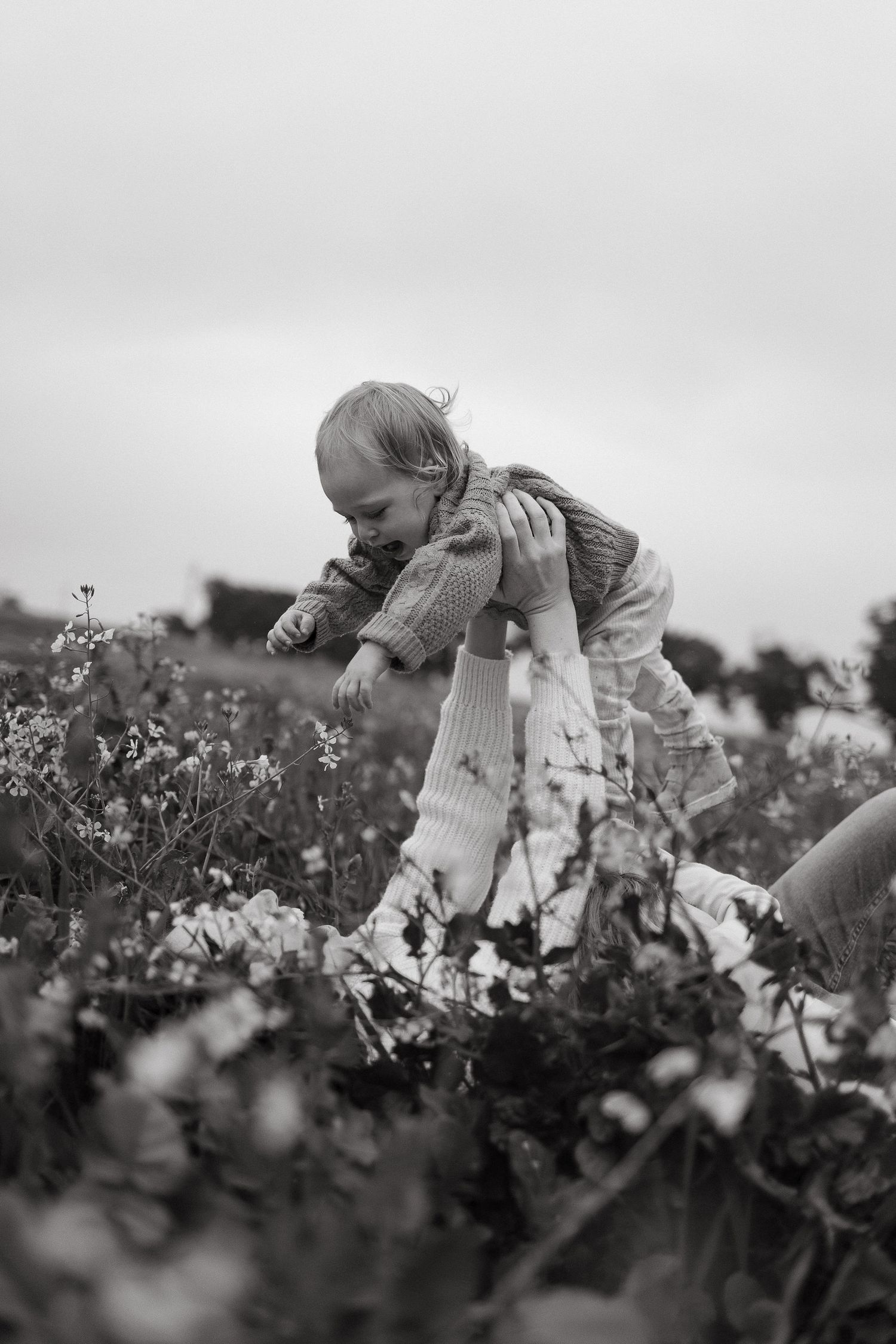 Black and white photo of a parent lifting a laughing toddler into the air above a field of wildflowers.