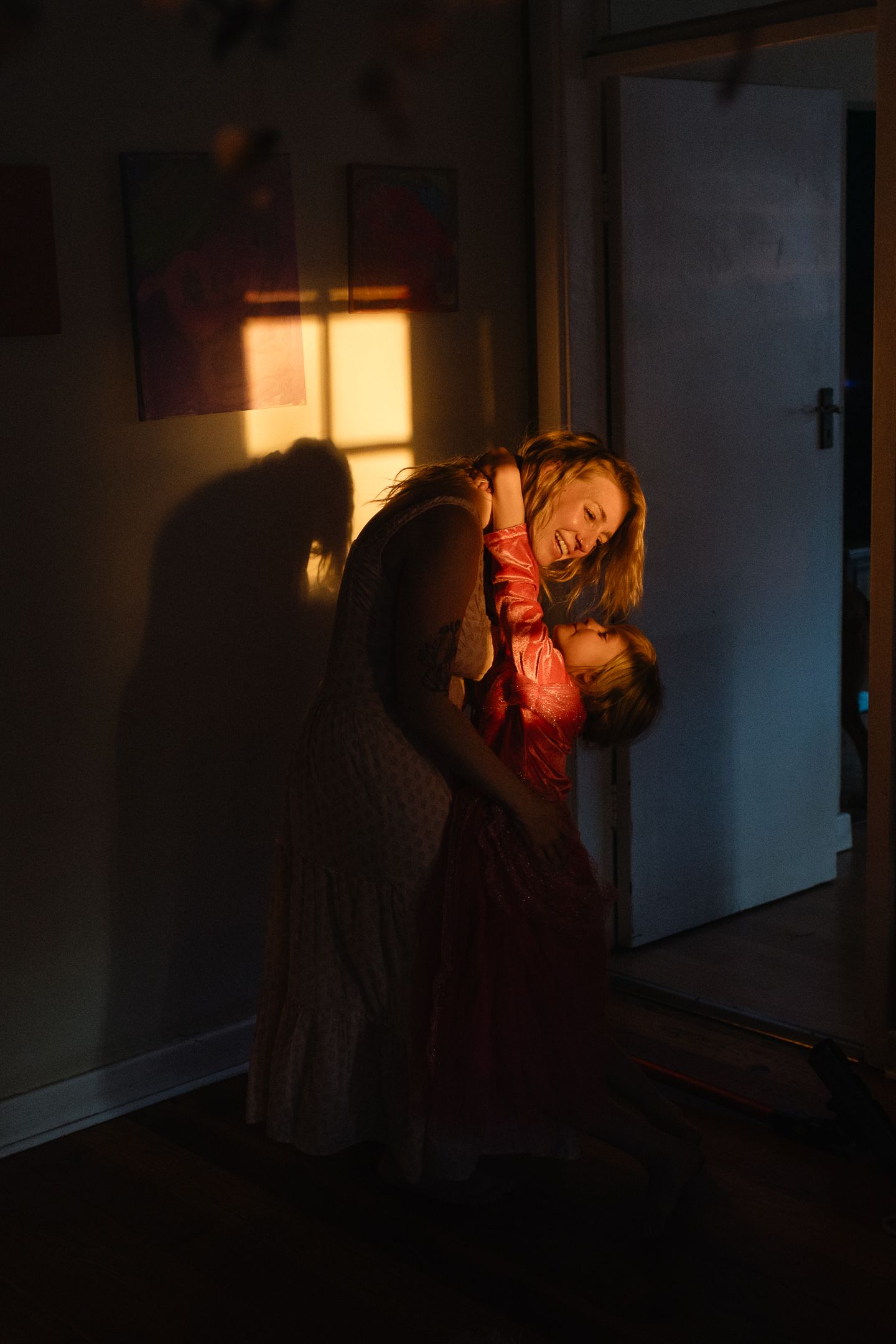 Mother lifts and embraces her laughing child in a dim room as warm golden light from a window casts glowing patches on the wall.