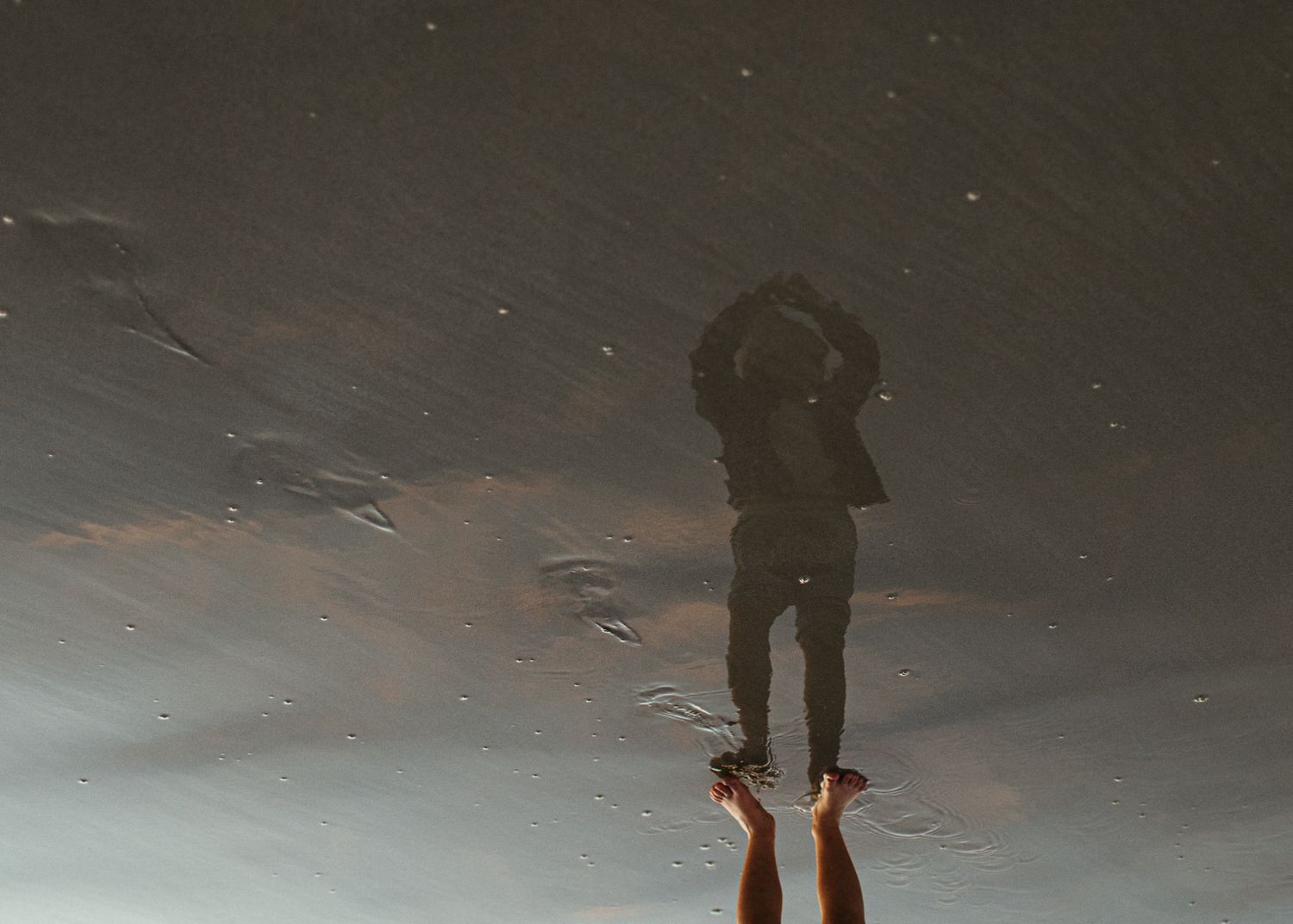 An upside-down reflection in wet sand shows a person's legs and silhouette, creating an abstract beach scene at dusk.