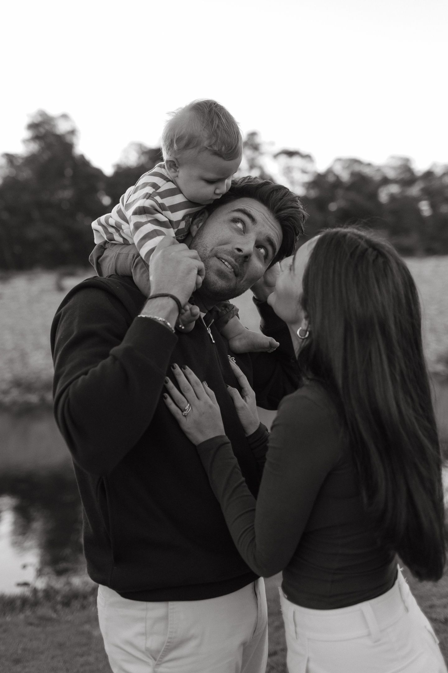Black and white photo of a father with a baby on his shoulders as a mother leans in to kiss him by a pond.