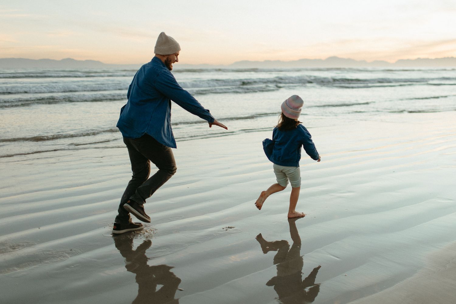 Parent and child in beanies run along the wet sand at sunset, their reflections mirrored in the shallow water.