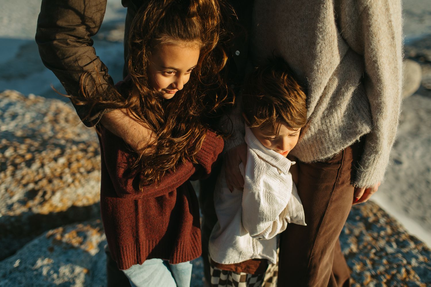 Two children huddle close to an adult in a cozy knit sweater while standing on a sunlit rocky beach.