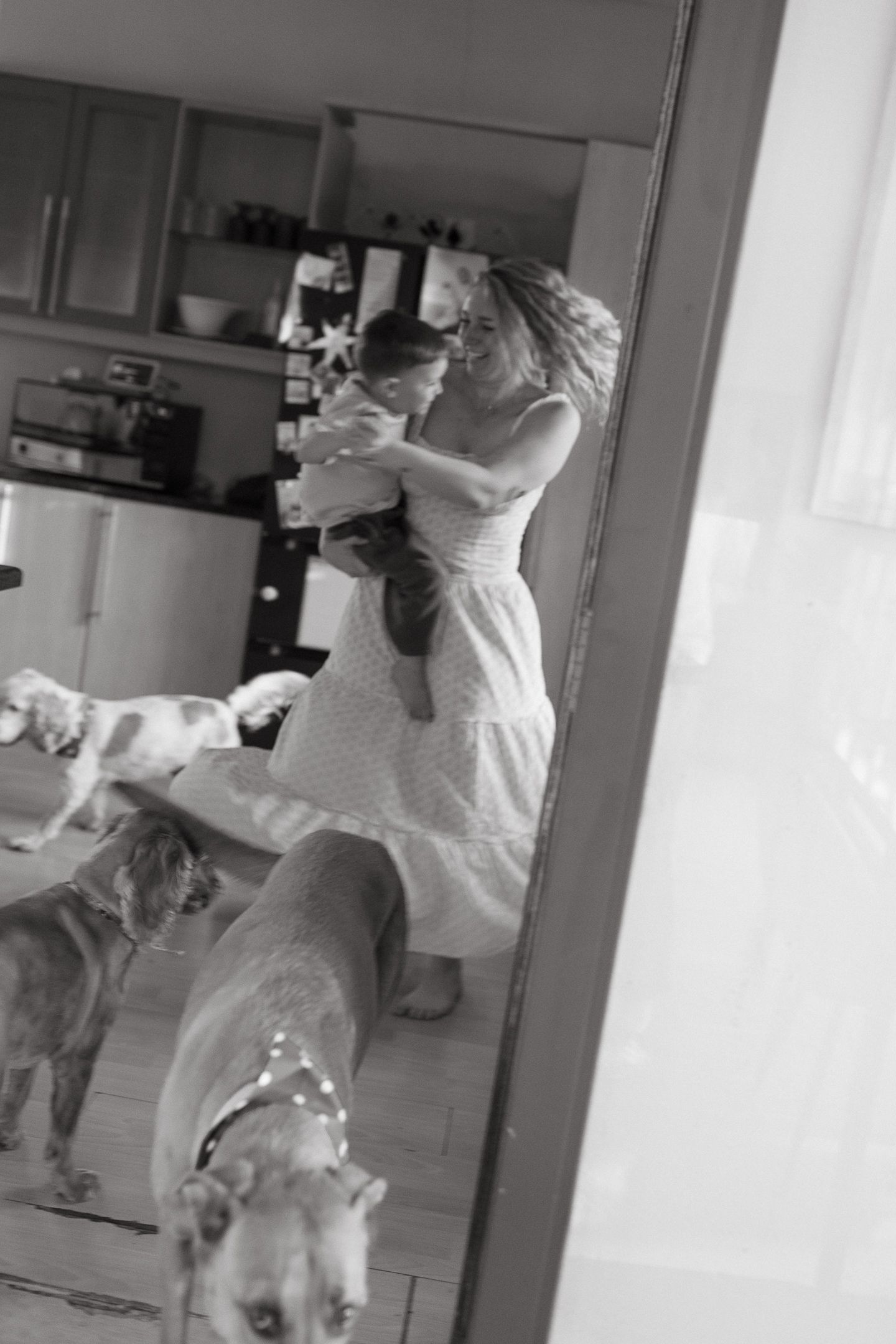 Black and white mirror reflection of a mother in a flowing dress holding a child while three dogs stand nearby.