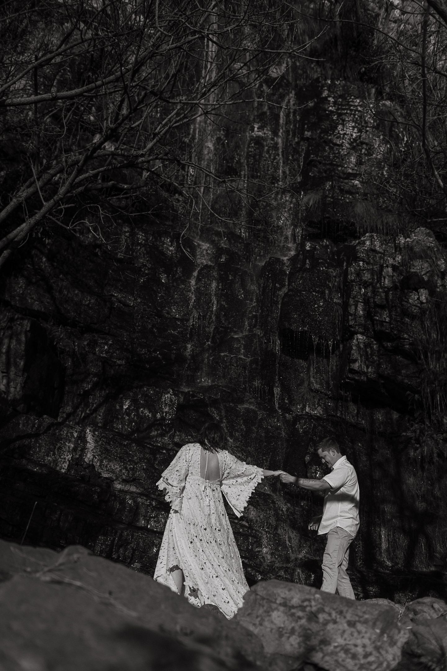 Black and white photo of a couple holding hands on rocks before a dark cliff, the woman in a flowing white lace dress.