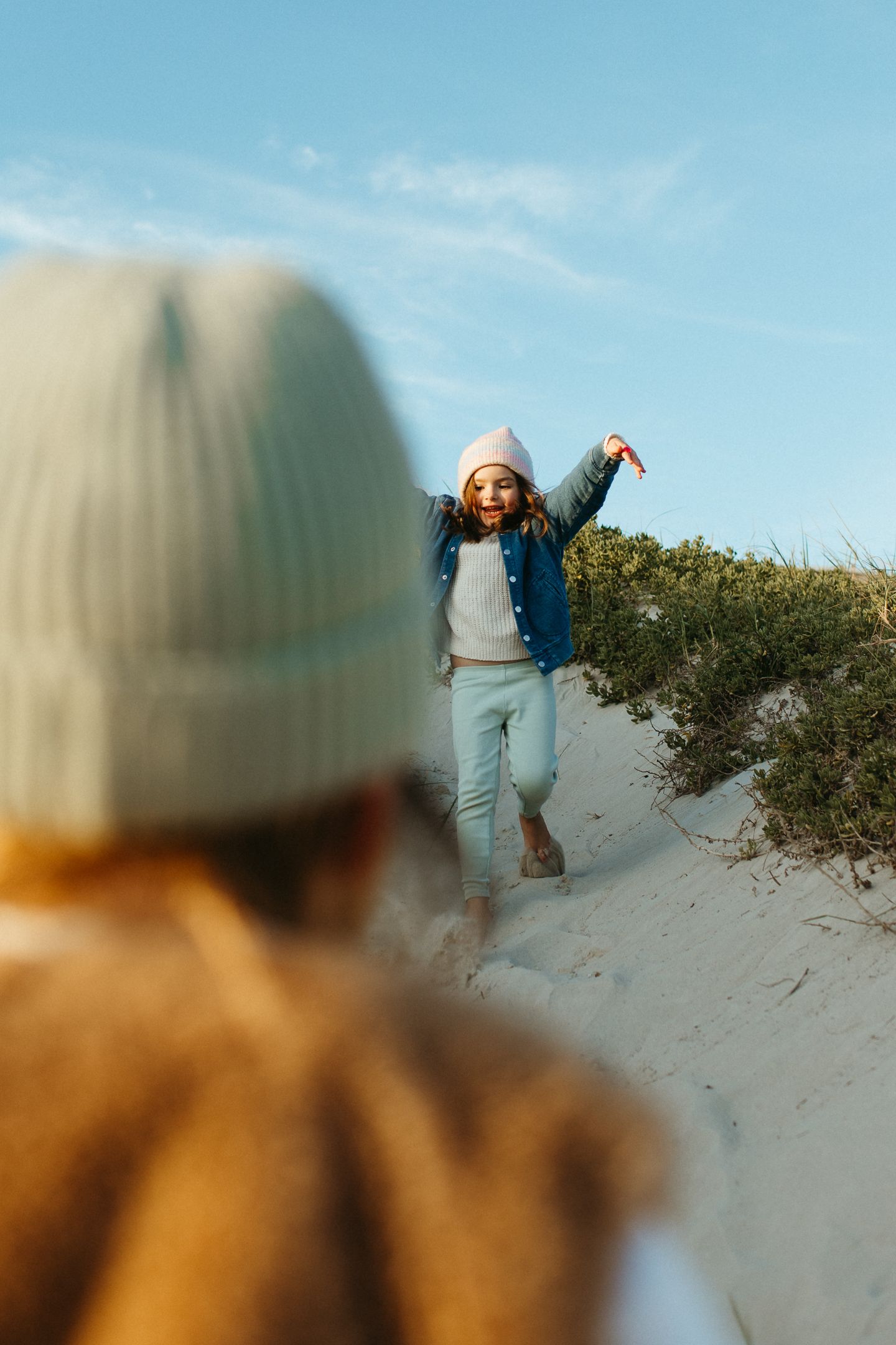 A smiling girl in a pink beanie and denim jacket runs down a sandy dune toward a blurred figure in a knit hat.