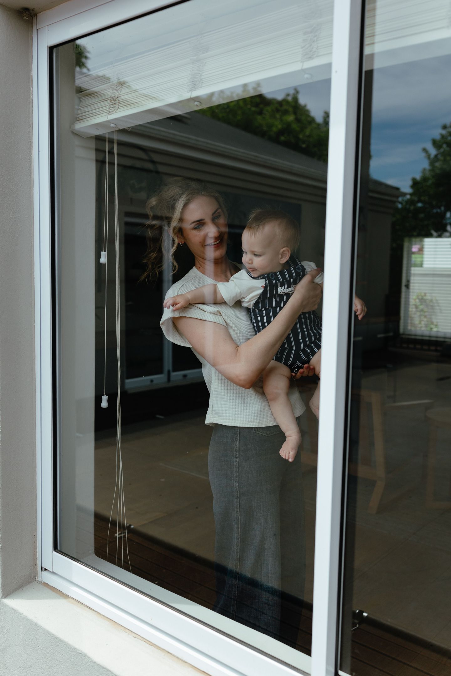 A smiling mother holds her baby in striped overalls, seen through a window with trees reflected in the glass.