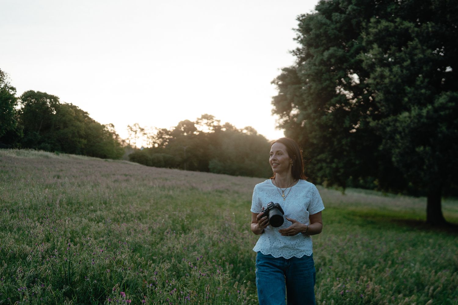 A woman holding a camera walks through a grassy meadow at dusk with tall trees and soft evening light behind her.