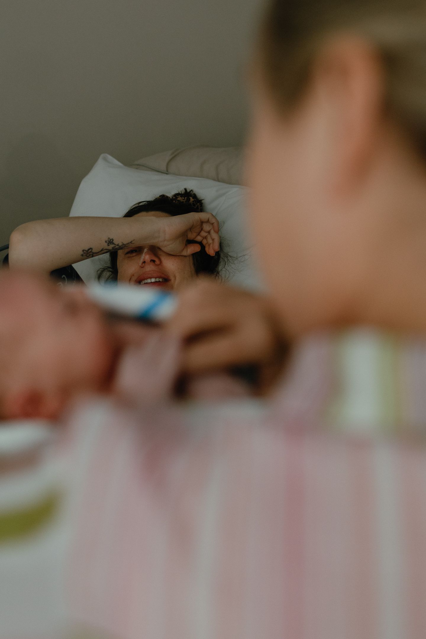 Smiling woman with a tattooed arm resting on her forehead lies in bed while a blurred newborn baby is held in the foreground.