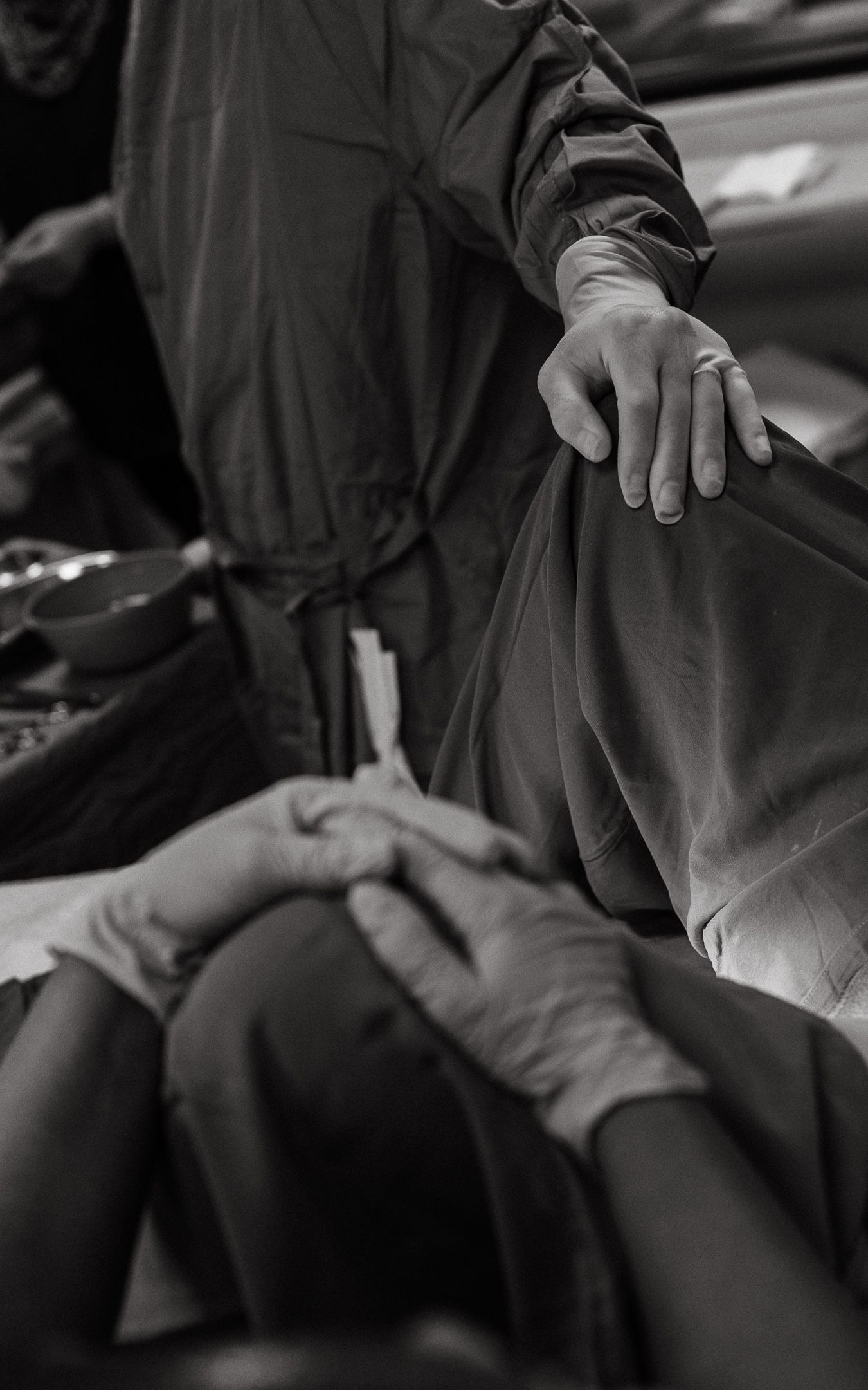 Black and white photo of a surgeon in a gown resting a hand on a patient's knee while gloved hands work in an operating room.
