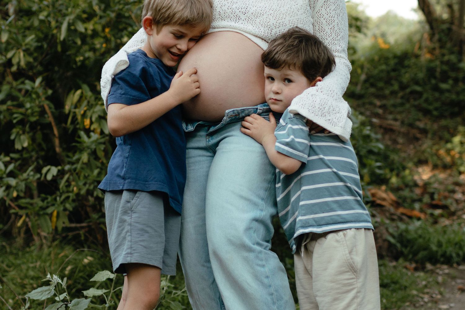 Two young brothers hug their pregnant mother's belly among green foliage during an outdoor maternity photo session.