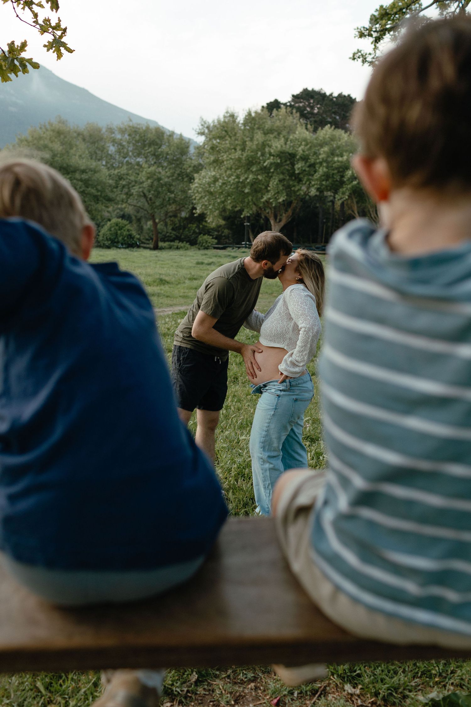A couple kisses while the woman holds her pregnant belly in a grassy park as two children sit on a bench in the foreground.
