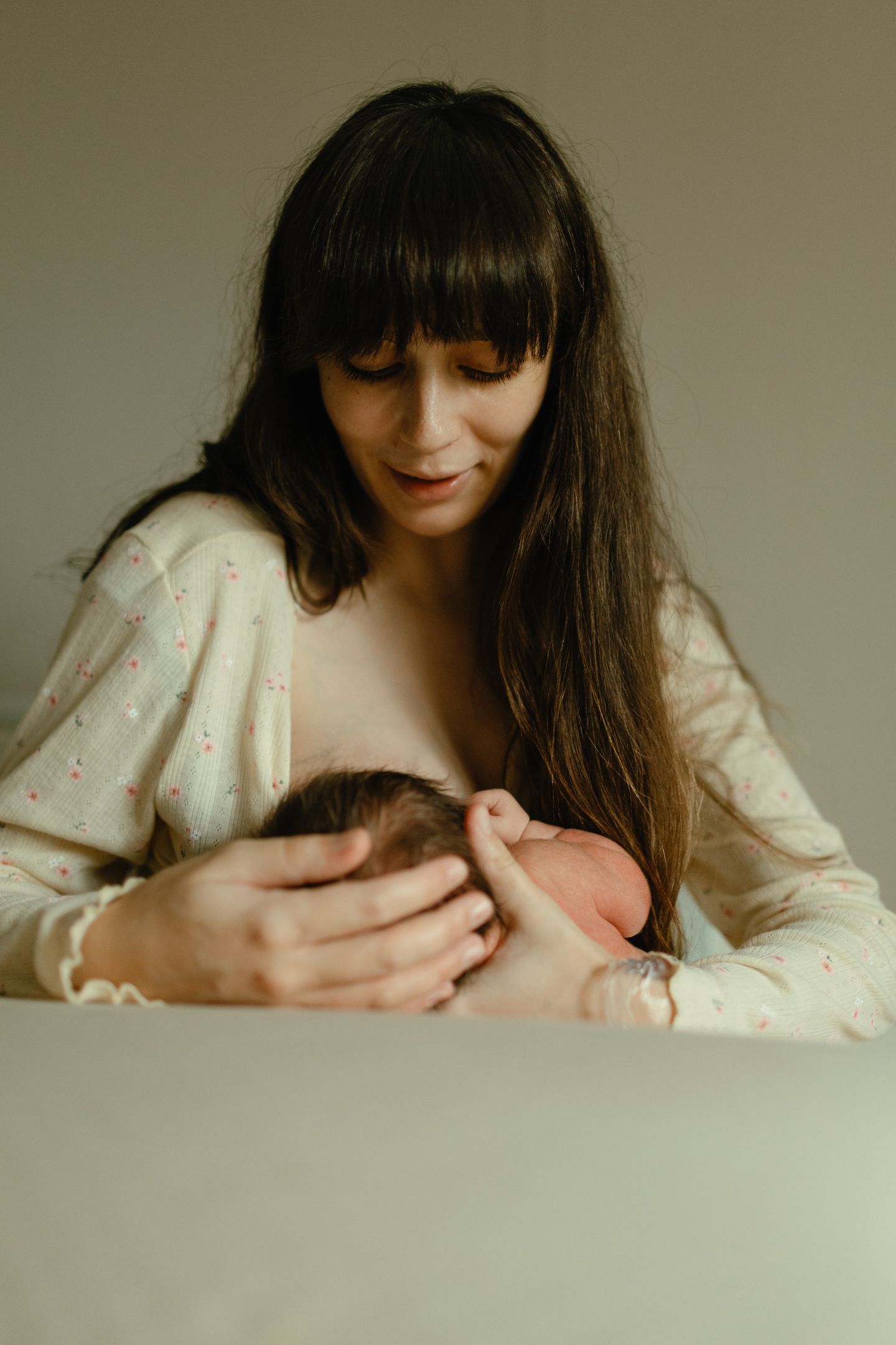 Mother with long dark hair and bangs breastfeeds her newborn baby while gently supporting its head.