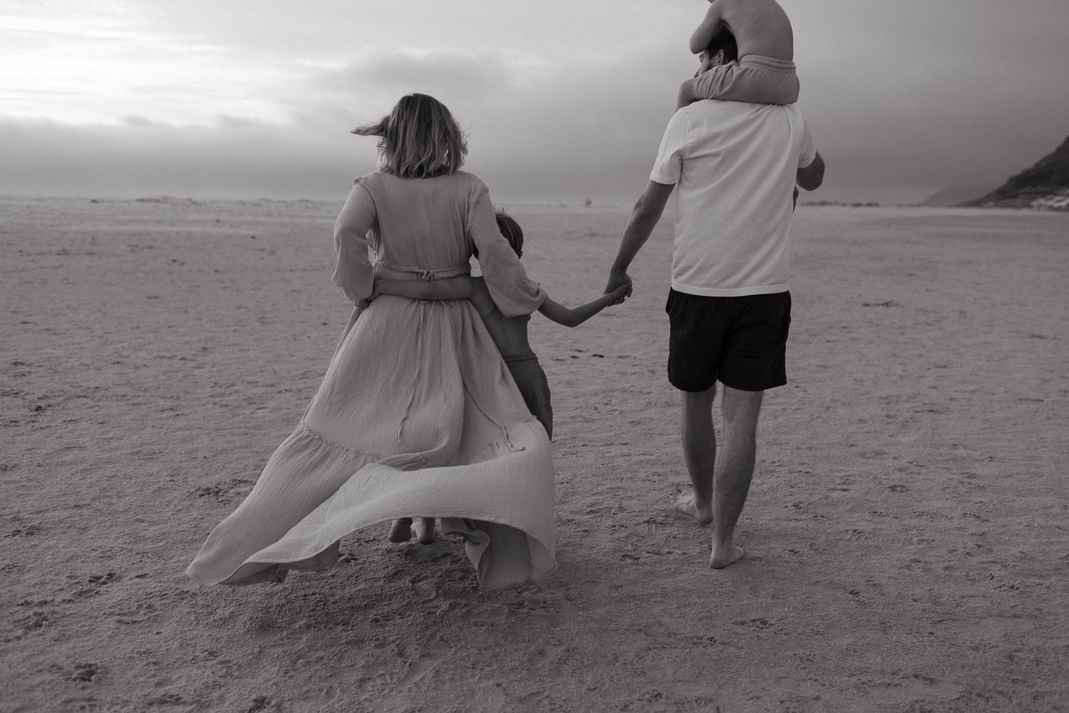 Black and white photo of a mother in a flowing dress and father holding hands with a child while walking on a beach.
