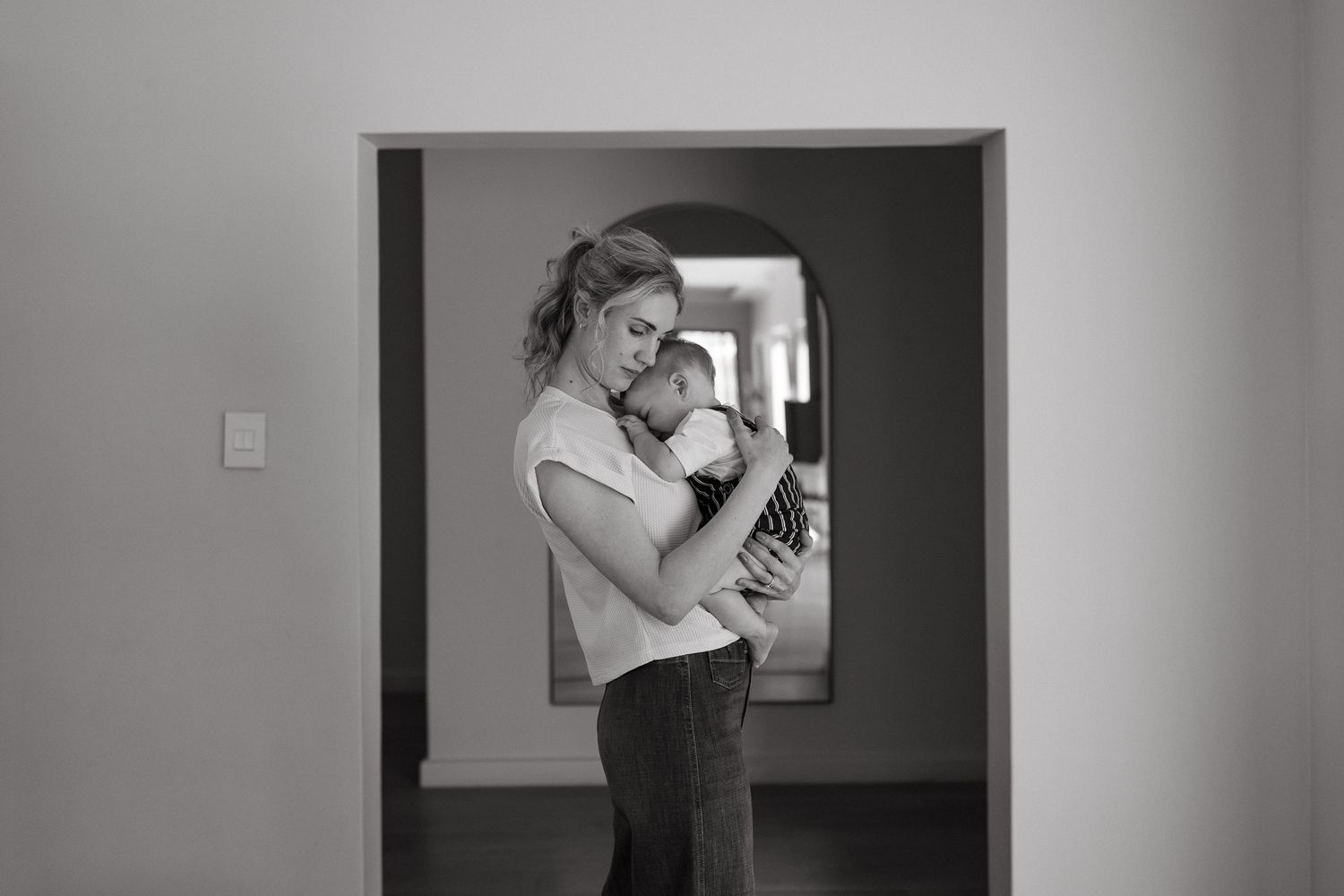 Black and white image of a mother holding her baby while standing in a doorway at home.