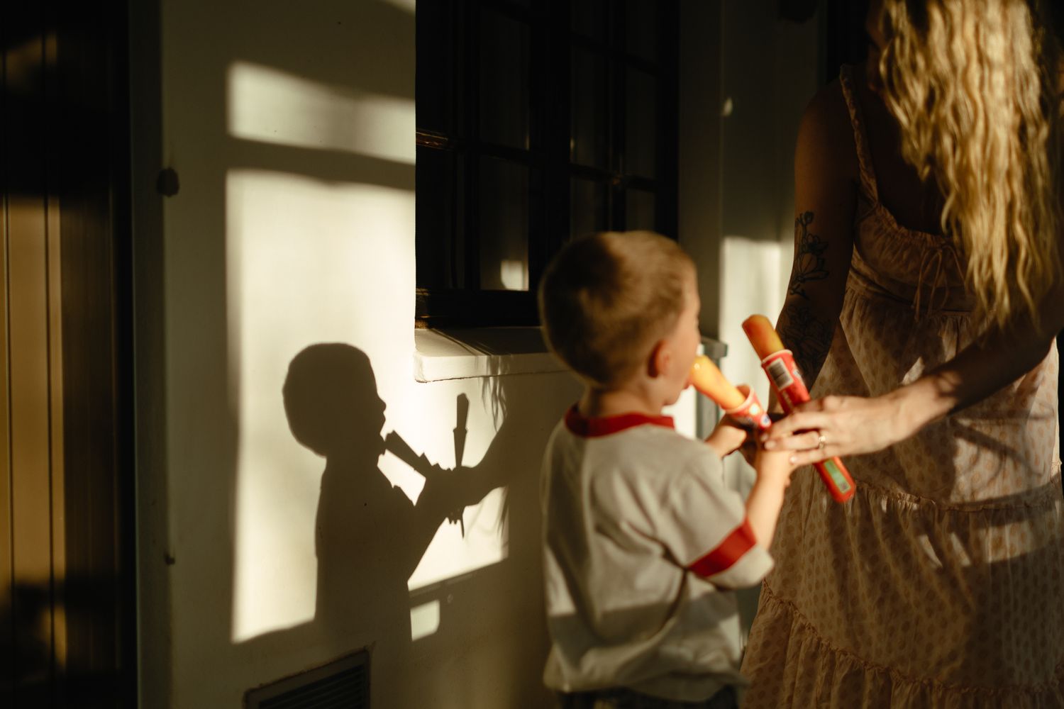 Warm golden light casts shadows on a wall as a little boy in a red-trimmed shirt plays recorders beside his mother in a sunlit room.