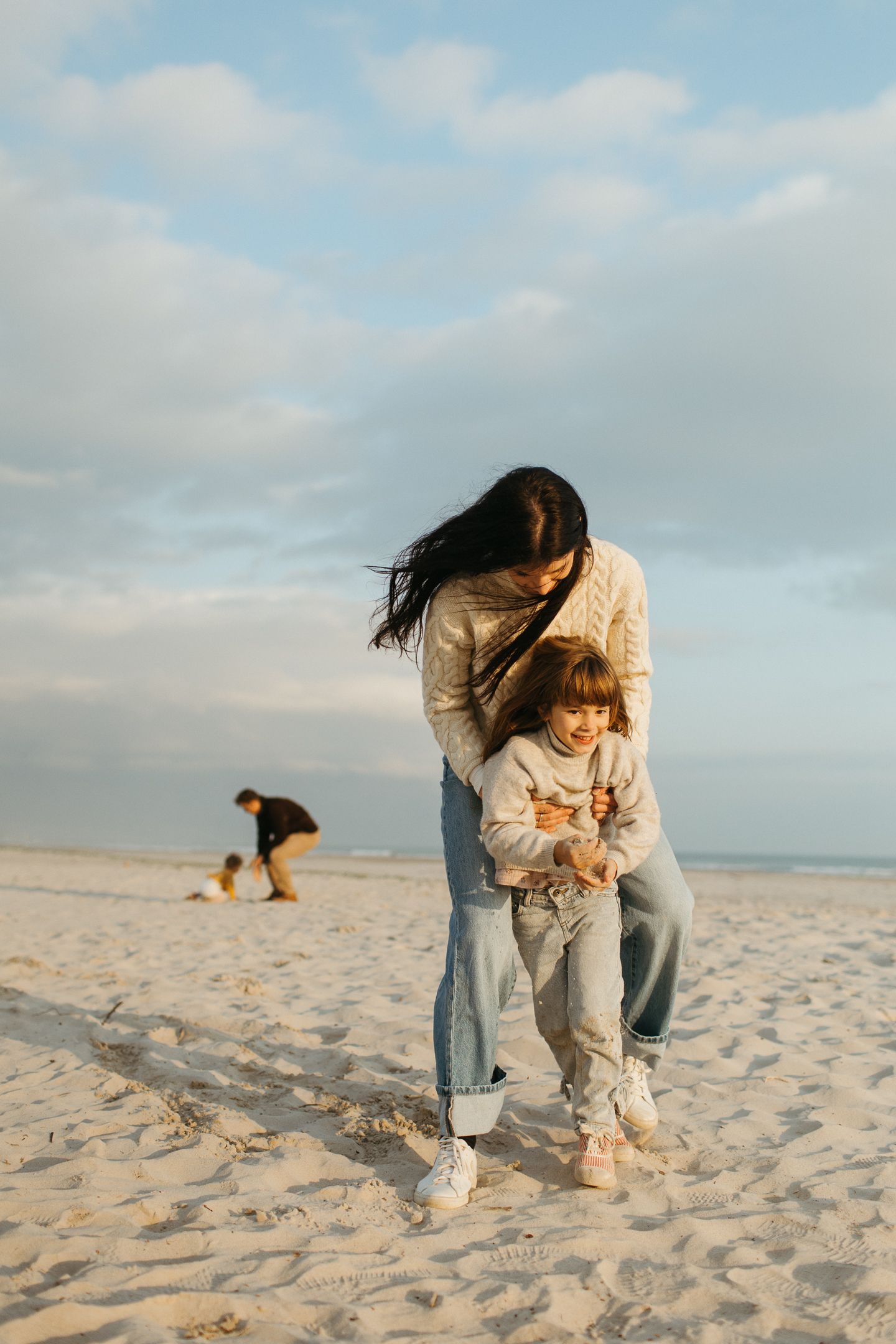 A dark-haired mother hugs her laughing toddler from behind on a wide sandy beach under a soft cloudy sky.
