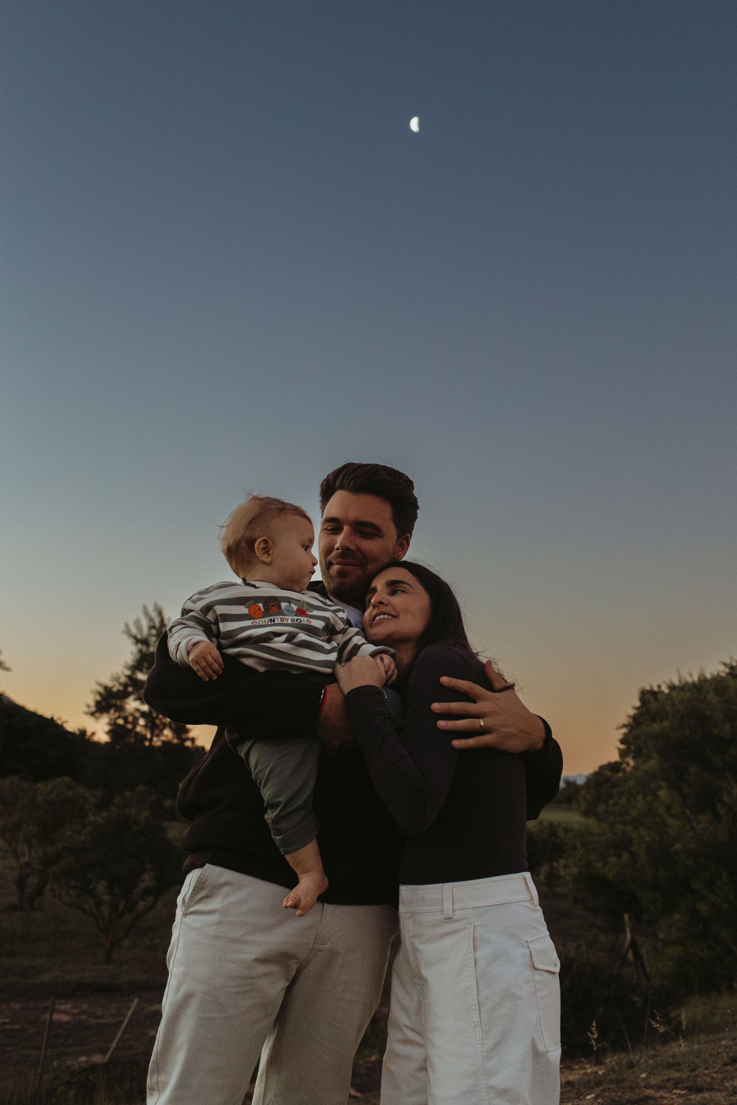 A smiling mother and father embrace their baby under a twilight sky with a crescent moon rising above the trees.