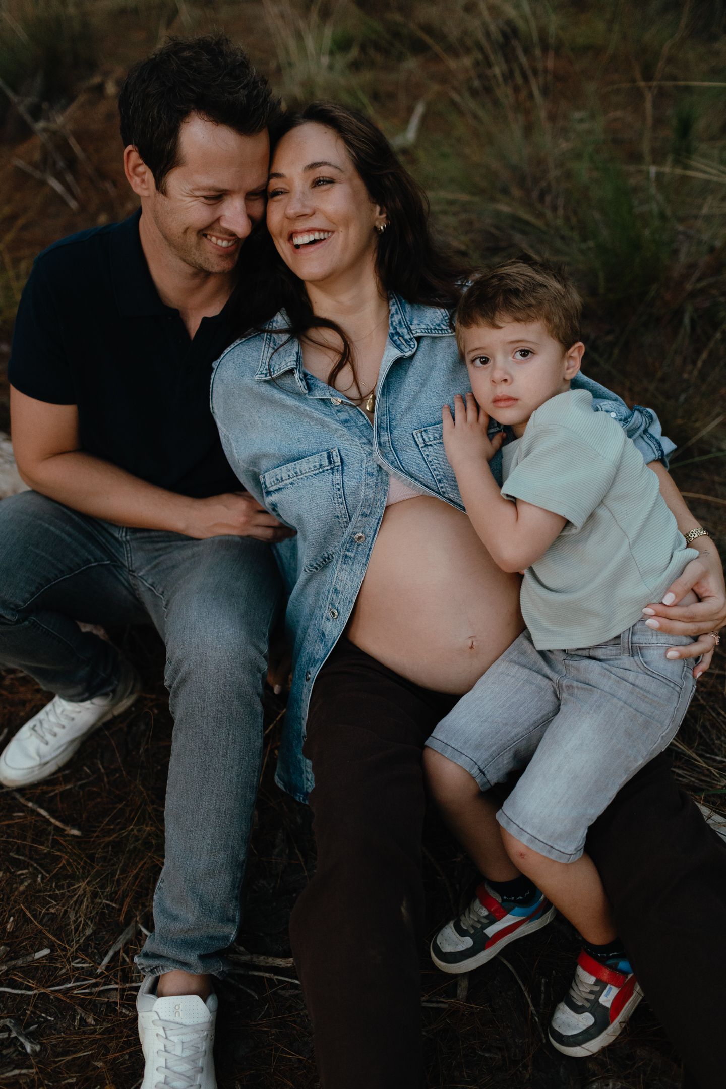Smiling couple sits on the ground in nature with a young boy, the pregnant woman in a denim jacket showing her baby bump.