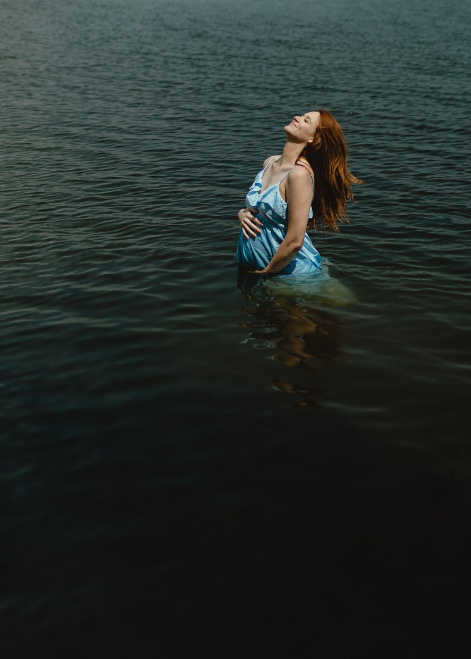 A pregnant woman in a flowing blue dress stands waist-deep in dark lake water with her head tilted back toward the sun.