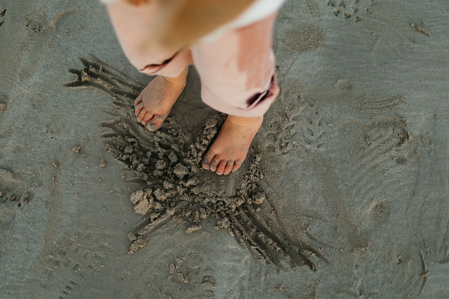 Child's bare feet standing in gray beach sand beside a small pile of sand sculpted into a star shape.
