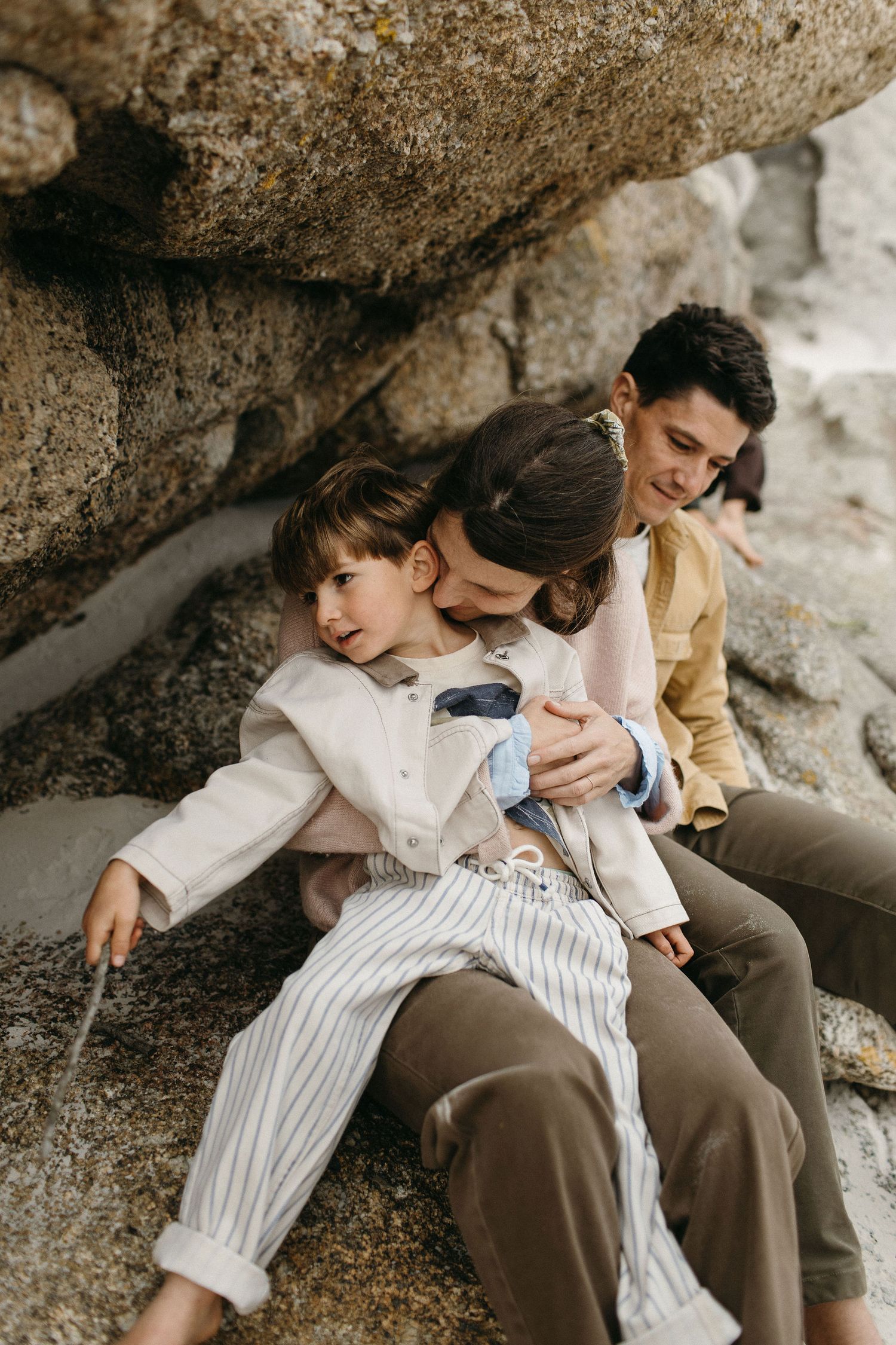 Mother cuddles a young boy in striped pants while the father smiles beside them on a rocky beach outcrop.