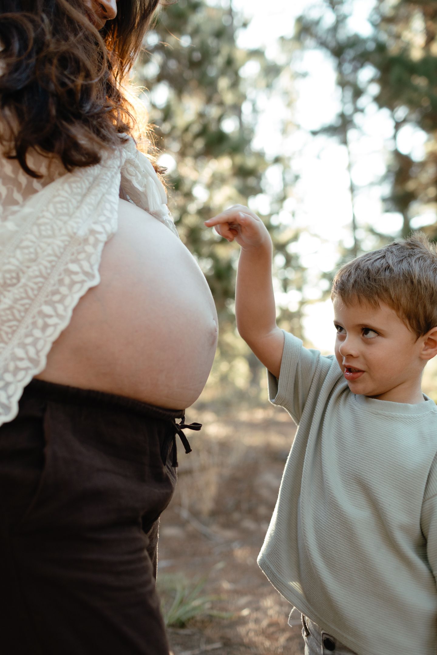 Toddler boy in a green shirt reaches out to touch his mother's pregnant belly in a sunlit forest.