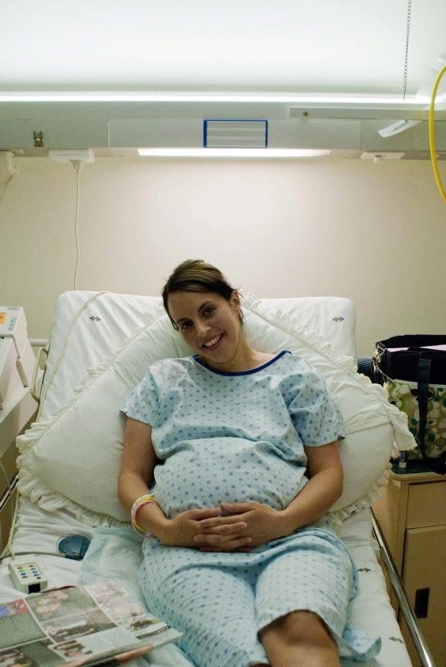 A smiling pregnant woman in a hospital gown rests her hands on her belly while sitting on a hospital bed.