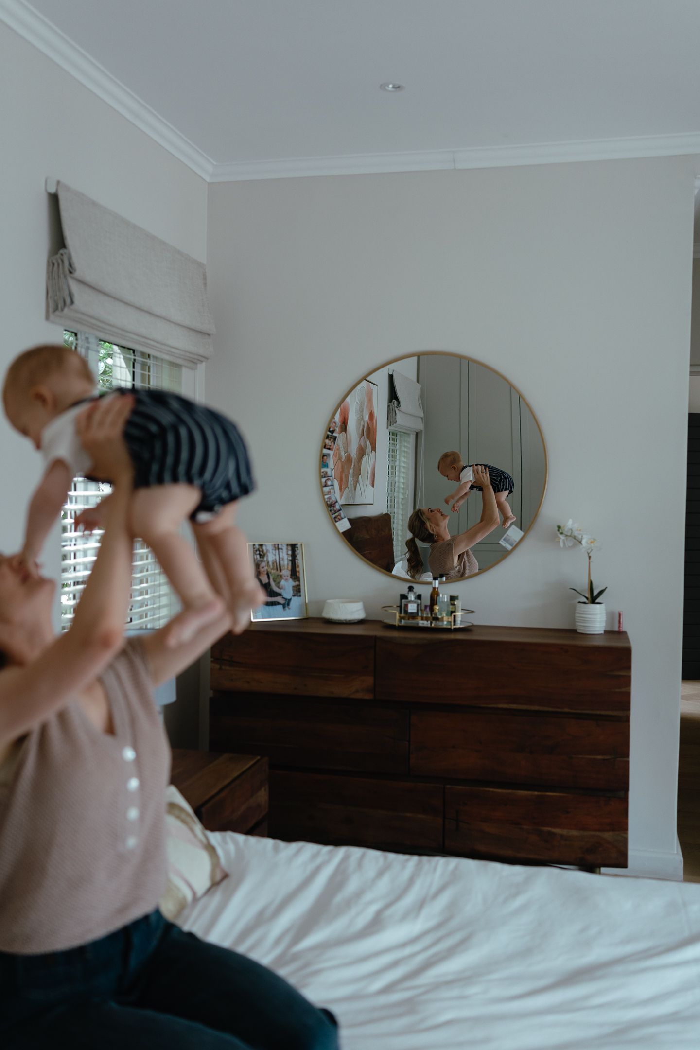 A mother lifts her baby overhead on a white bed, with the pair reflected in a round mirror above a wooden dresser.