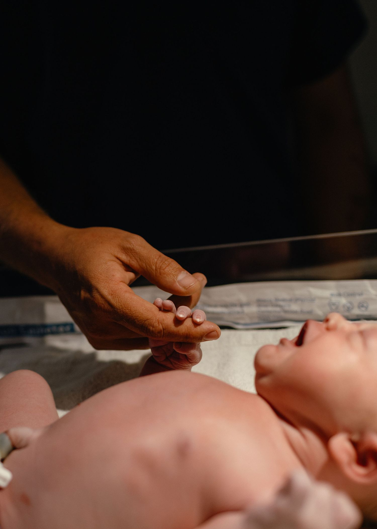 A parent's hand gently touches the fingers of a crying newborn lying on a hospital bassinet under warm light.