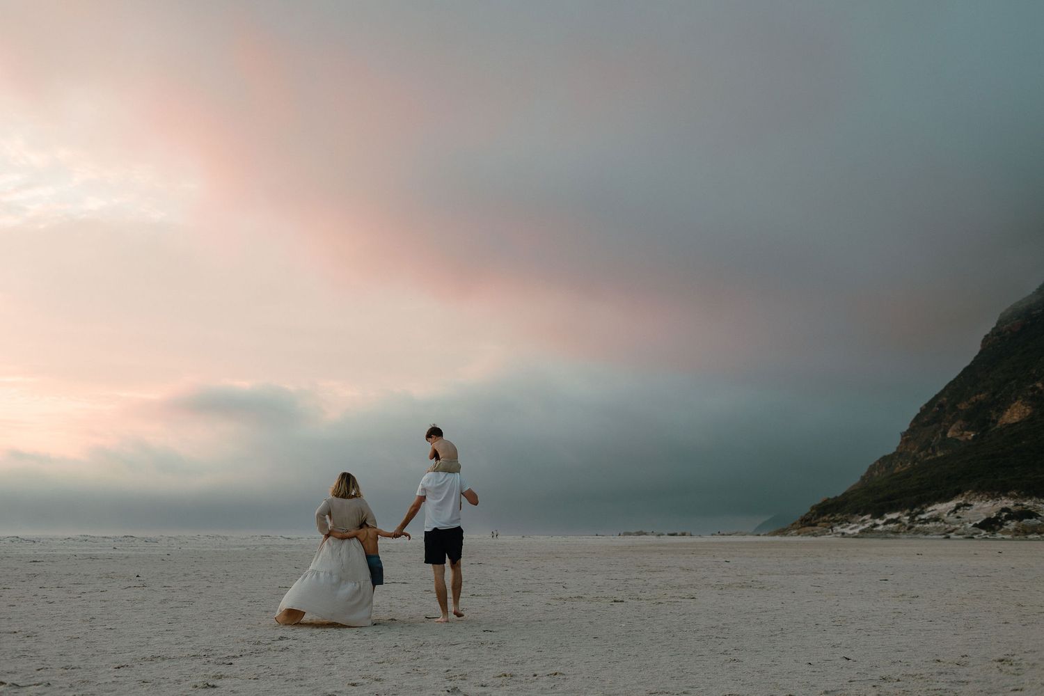 Couple in a flowing dress and white shirt hold hands while walking on a sandy beach beneath a pink and gray sunset sky.