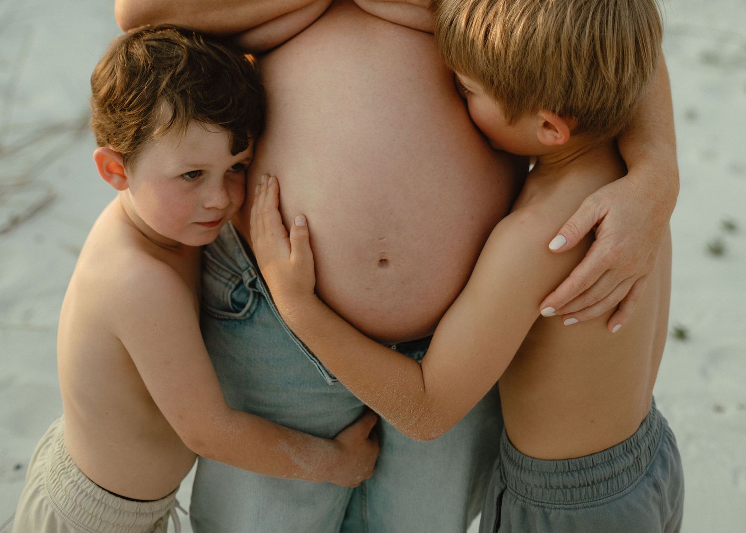 Two shirtless boys hug their pregnant mother's bare belly as she stands on a beach in jeans.