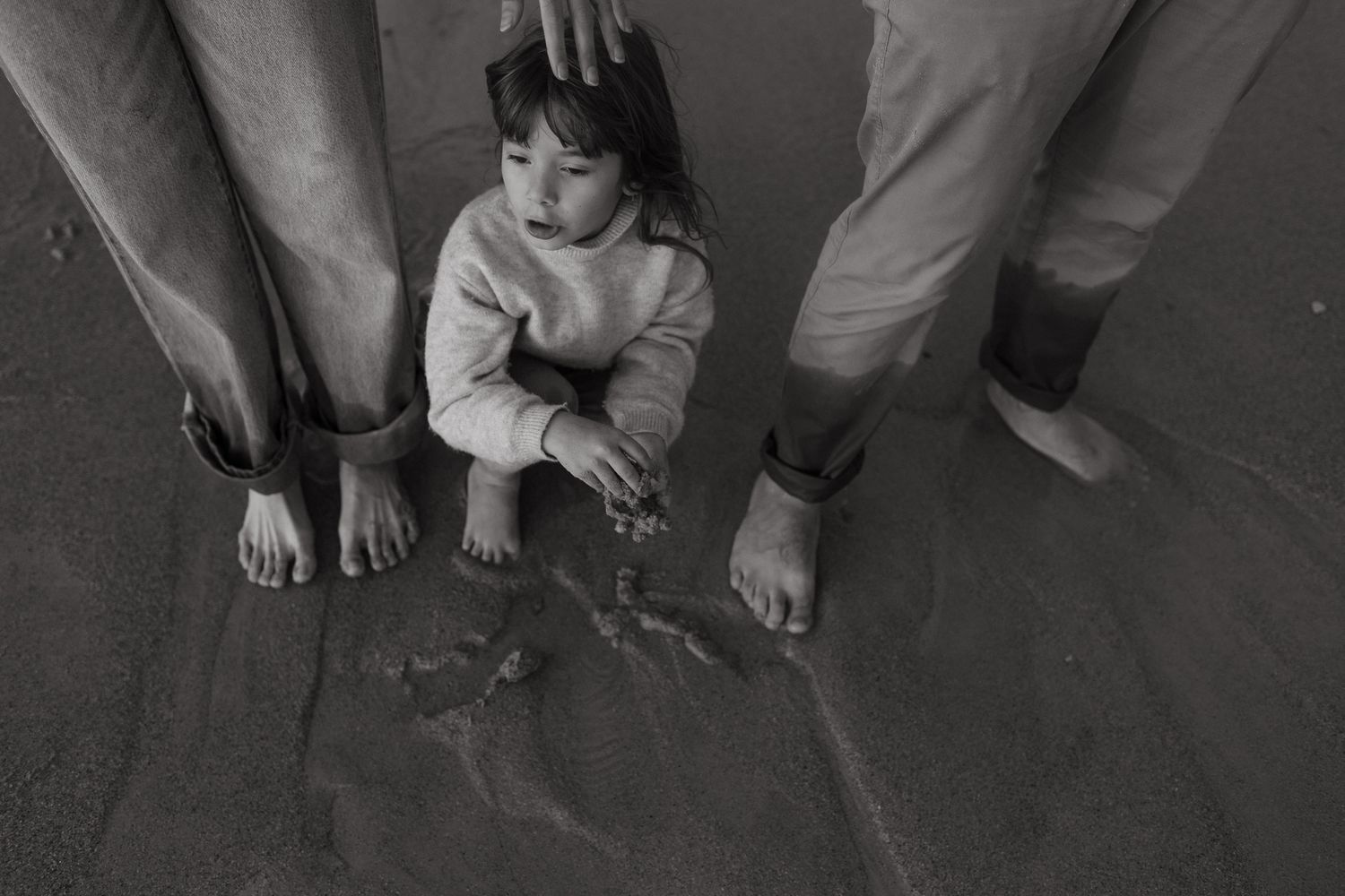 Black and white overhead view of a young child standing barefoot on sand between two adults' legs.