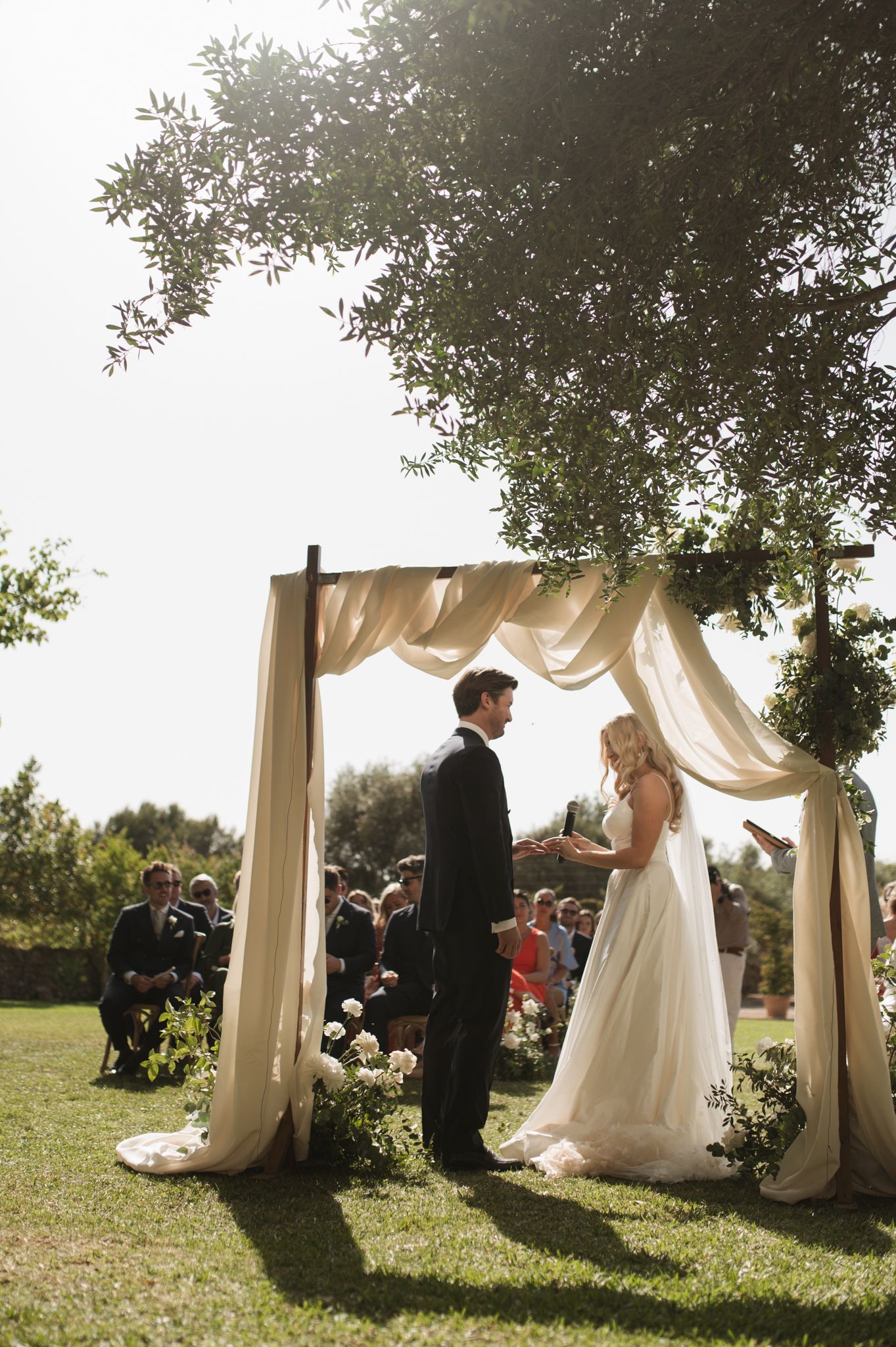 Bride and groom exchange vows under a draped fabric wedding arch surrounded by lush greenery outdoors.