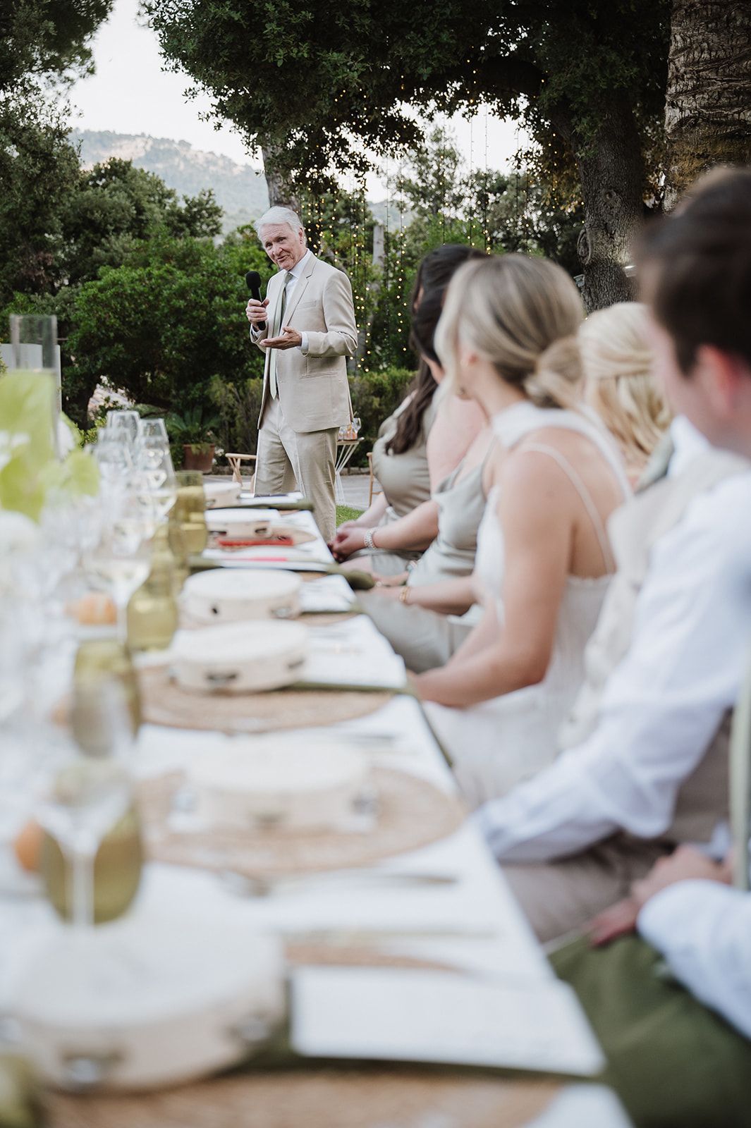 Elegant outdoor wedding reception with guests seated at long table while man in suit gives a toast.