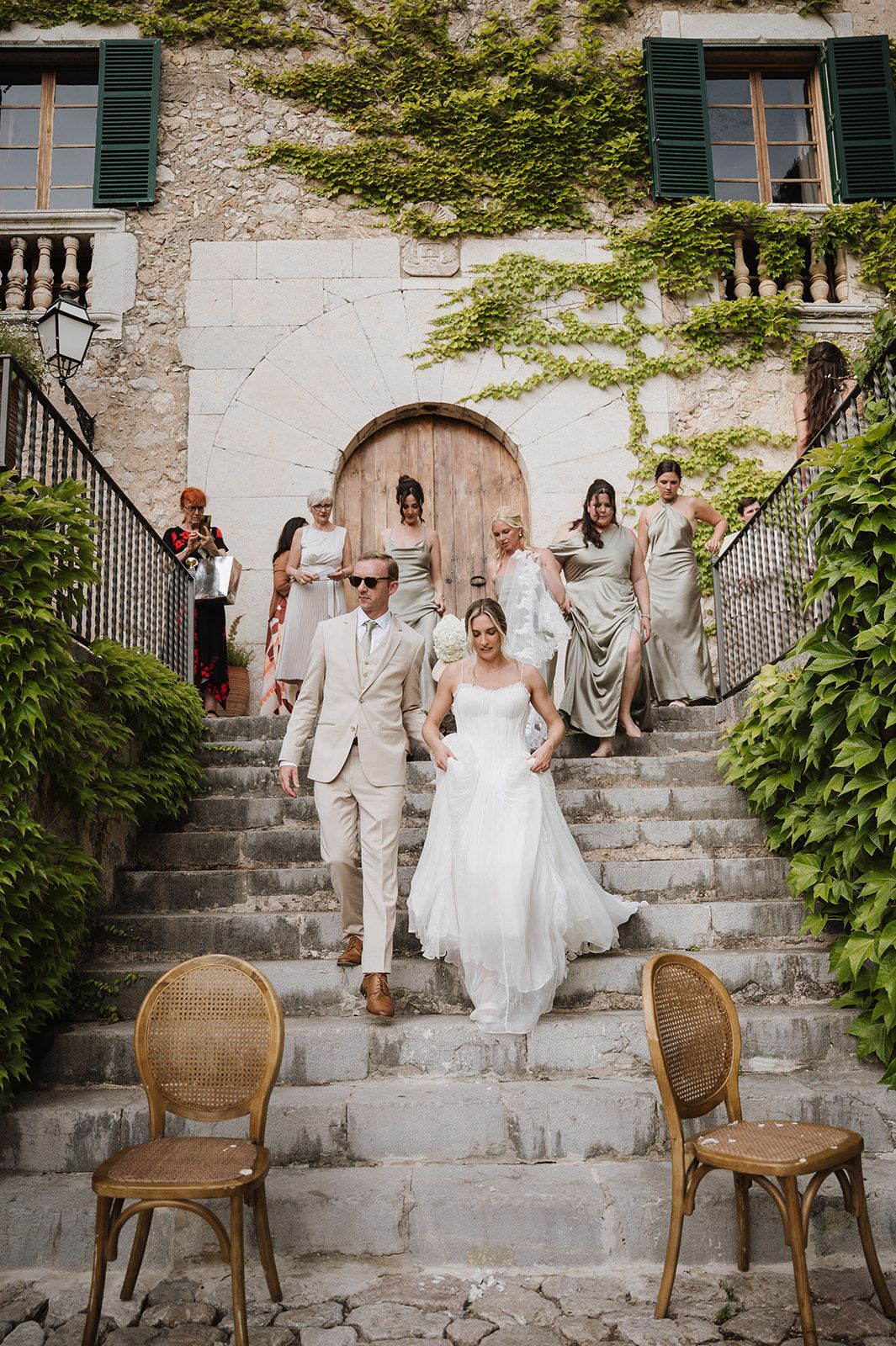 Bride and groom descend stone steps of ivy-covered villa with wedding party following, bridesmaids in sage green gowns.