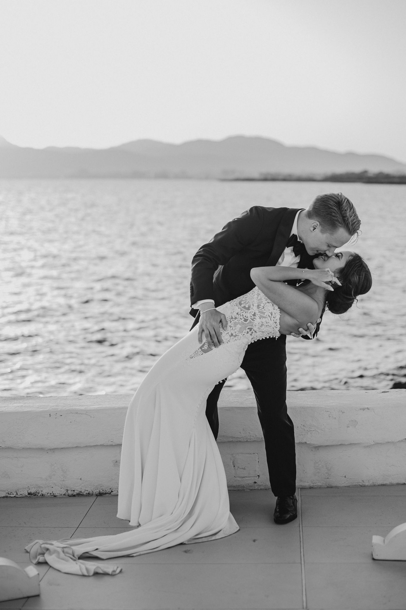 Black and white romantic photo of groom dipping bride for a kiss by the ocean at sunset after wedding.