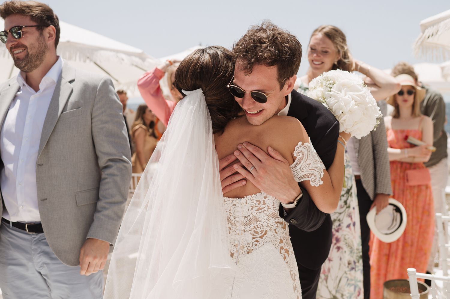 Groom embraces laughing bride in lace wedding dress during outdoor Mediterranean wedding reception.
