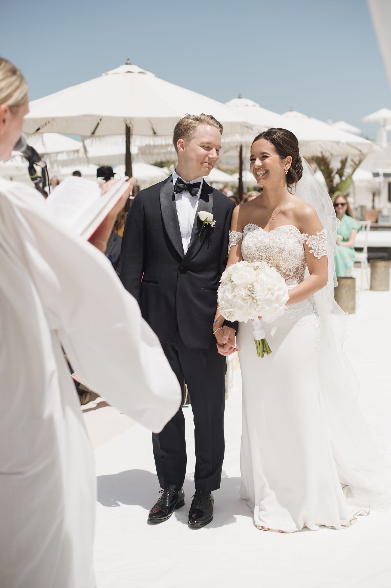 Smiling bride and groom holding hands during outdoor beach wedding ceremony with white umbrellas in background.