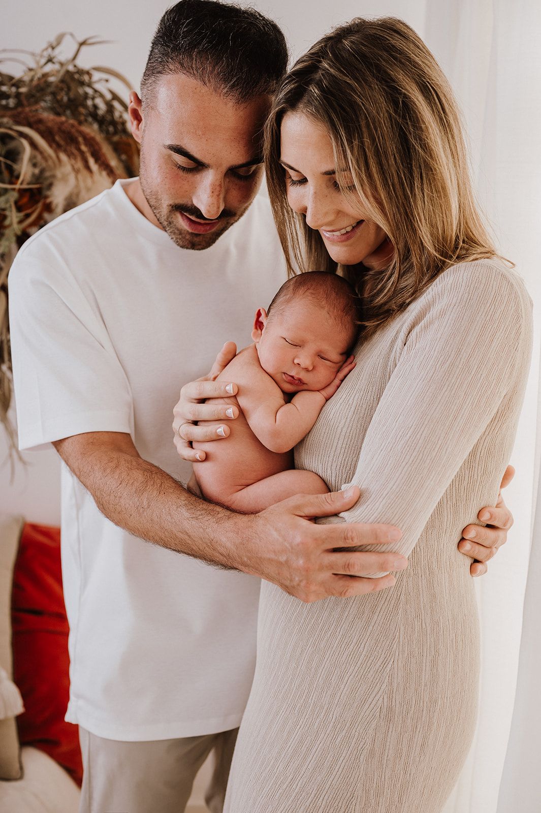 Happy parents in white outfits tenderly hold their sleeping newborn baby together in a bright, warm room.
