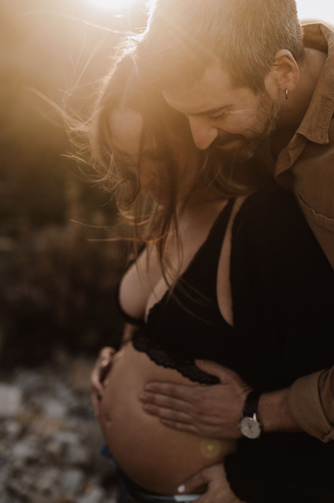 A bearded man tenderly embraces a pregnant woman from behind in golden sunset light, both gazing lovingly at her baby bump.