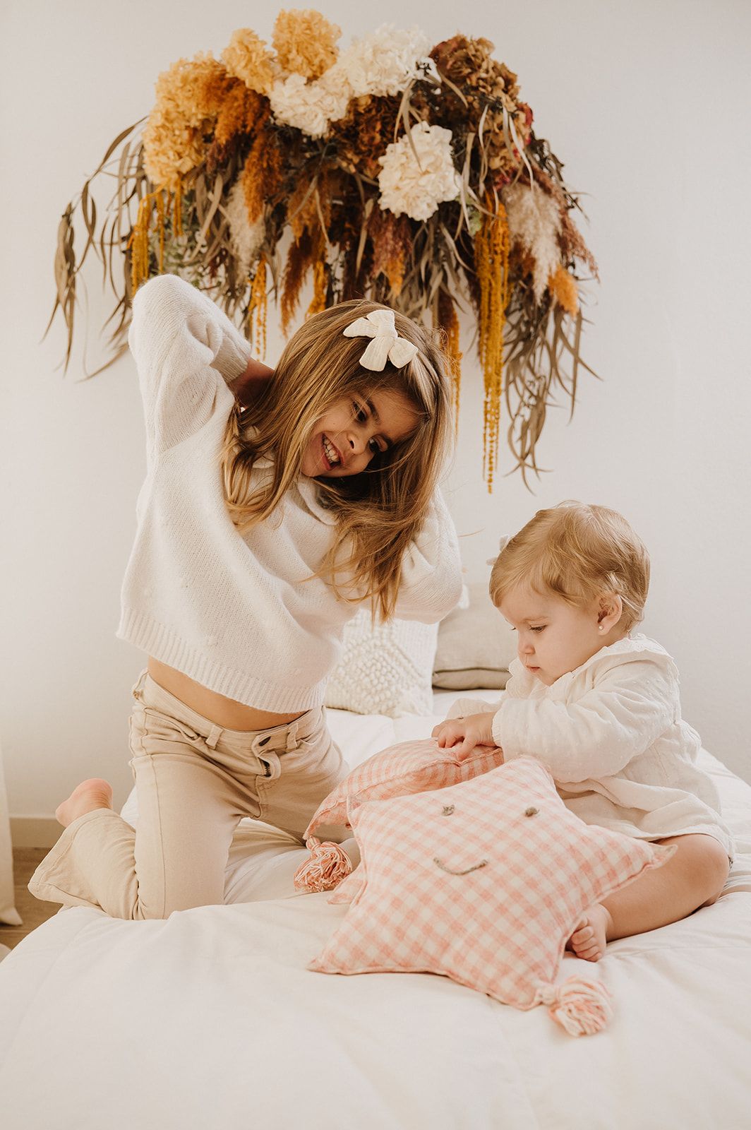 Mother and toddler playing with pink gingham pillow on white bed under dried florals.